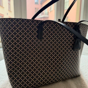 Svart mönstrad handväska By Malene Birger - Stilren handväska från By Malene Birger med svart och beige rutigt mönster. Väskan har två svarta handtag och rymligt innandöme med dragkedjeficka. Materialet är syntet med detaljer i skinn. Perfekt för dig som gillar klassisk design med modern twist. Nypris 2800kr. Storlek 48 x 28 x 12.5cm.