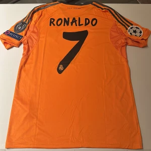 Real Madrid Ronaldo 7 orange matchtröja - Säljer en orange Real Madrid fotbollströja med Ronaldo och nummer 7 på ryggen. Tröjan har svarta detaljer, Champions League-patchar på ärmarna och klassiska Adidas-ränder. Tillverkad i lätt och ventilerande polyester, perfekt för fotboll eller samling. Replika, endast handtvätt gäller!
