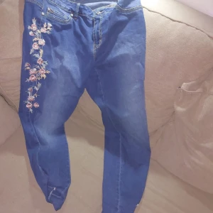 Blå jeans med broderade blommor - Snygga blå jeans från bonprix i storlek 42 med broderade rosa och vita blommor längs ena benet. Klassisk femficksmodell med dragkedja och knapp. Jeansen har råa, fransiga benslut och är tillverkade i mjukt bomullsjeansmaterial.