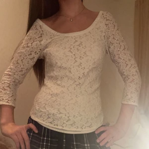 Vit spetsblus med pärlknappar - Superfin vit blus i spets med blommigt mönster och pärlknappar längs hela framsidan. Blusen har v-ringning, trekvartsärmar och kan bäras både med knapparna fram eller bak för olika look. Perfekt för dig som gillar romantiska detaljer och unik stil.