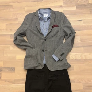 Ullkavaj J.Lindeberg - Grå kavaj från J.Lindeberg i fiskbensmönstrad ullmix. Klassisk passform med patchfickor fram, kissing buttons vid ärmslut och armbågsskydd – perfekt för resor. Ofodrad för en lättare känsla.  Mått (cm): 	•	Armhåla till armhåla: 48 	•	Längd (nacke till botten): 74 	•	Ärmlängd: 63 	•	Axelbredd: 44