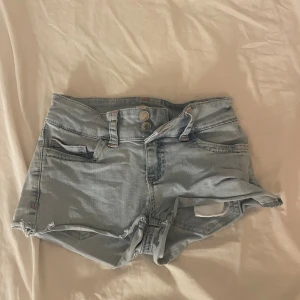 Ljusblå jeansshorts från Nelly - Säljer ett par ljusblå jeansshorts från Nelly med hög midja, råa kanter och dubbla knappar framtill. Shortsen har klassiska fickor och en bakficka med orange broderad detalj. Perfekta för varma dagar och en avslappnad look. Vet inte vilken storlek men det är den minsta Nelly har💕💖💓
