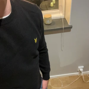 Svart stickad tröja från Lyle & Scott - Svart stickad tröja från Lyle & Scott med klassisk rund halsringning och gul broderad logga på bröstet. Tröjan har lång ärm och ribbade muddar vid ärmslut och nederkant. Perfekt för lager-på-lager och snygg till jeans.
