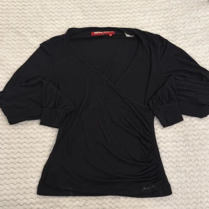 Svart omlottopp från Miss Sixty - Svart omlottopp från Miss Sixty med v-ringning och trekvartsärmar med breda muddar. Toppen har en figurnära passform och är tillverkad i mjukt material. Diskret logga broderad nertill framtill. Perfekt för en trendig och stilren look.