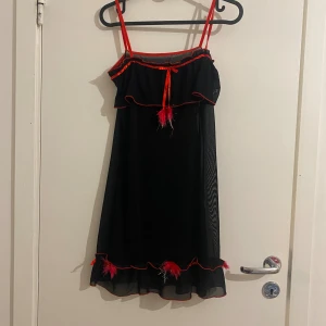 Svart nattlinne  - Svart kort nattlinne meshklänning med tunna röda axelband och röda volangkanter. Klänningen har fjäderdetaljer framtill och nertill samt transparent tyg som ger en edgy vibe. Perfekt för dig som gillar unika och lekfulla plagg.