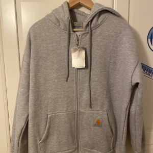 Grå hoodie med dragkedja från Carhartt (HELT NY) - Snygg ljusgrå hoodie från Carhartt med dragkedja, huva och stora fickor framtill. Klassisk Carhartt-logga på fickan och justerbar snörning i huvan. Perfekt för chill dagar och streetwear-vibe.