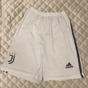 Juventus vita Adidas shorts - Snygga vita Juventus shorts från Adidas med svarta ränder längs sidorna och klubbens logga med tre stjärnor broderad på ena benet. Elastisk midja för skön passform, perfekt för fotboll eller träning. Tillverkade i lätt och ventilerande polyester.