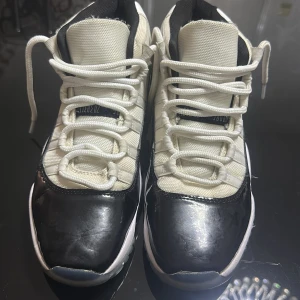 Nike Air Jordan 11 Concord Highs - Säljer ett par Nike Air Jordan 11 Concord med högt skaft. Skorna är vita och svarta med tydliga Jumpman-loggor och siffran 45 på hälen. De har snörning, rund tå och en blandning av textil och glansigt syntetmaterial. Perfekta för dig som gillar klassiska sneakers med ikonisk design.