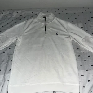 Vit half zip tröja från Polo Ralph Lauren - Vit långärmad tröja från Polo Ralph Lauren med klassisk broderad logga på bröstet. Tröjan har en snygg half zip-dragkedja vid halsen och är gjord i mjuk bomull. Perfekt för en clean och stilren look.