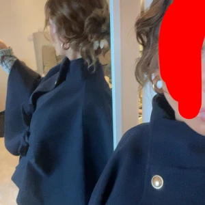 Mörkblå kappa med vida ärmar - Snygg mörkblå kappa med vida ärmar och hög krage. Kappan har dekorativa knappar framtill och en elegant, stilren design som ger en sofistikerad look. Perfekt för dig som vill ha något klassiskt men ändå trendigt. men knappen har då gått av men har användt den efter med att ha en säkerhetsnål på insidan av jackan så synns det ej 