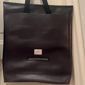 Eytys svart axelväska  - Stilren svart axelväska från Eytys i vattenavvisande syntetmaterial. Väskan har rejäla handtag, innerfack med dragkedja och diskret Eytys-logga framtill. Perfekt för dig som vill ha en minimalistisk och modern look.