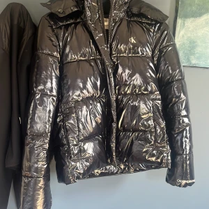 Svart glansig pufferjacka Calvin Klein - Svart pufferjacka från Calvin Klein med glansig finish och diskret CK-logga på bröstet. Jackan har hög krage, huva och dragkedja framtill. Fylld med syntetmaterial och har långa ärmar. Perfekt för kalla dagar när du vill ha en cool look.