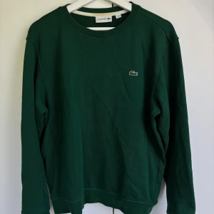 Grön sweatshirt från Lacoste - Mörkgrön sweatshirt från Lacoste med klassisk rund hals och det ikoniska krokodilmärket broderat på bröstet. Tröjan har långa ärmar och är tillverkad i mjuk bomull, perfekt för en chill och stilren look.