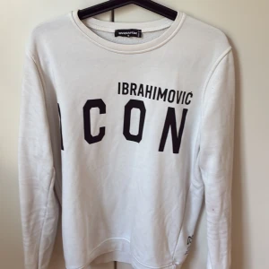 Vit sweatshirt Dsquared2 ICON Ibrahimovic - Vit sweatshirt från Dsquared2 med svart text 'IBRAHIMOVIC ICON' på både fram- och baksidan. Rund halsringning och långa ärmar. Mjuk bomullskvalitet och klassisk passform. Perfekt för dig som gillar sportiga och trendiga plagg med statement-tryck.