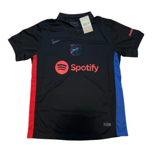 Svart FC Barcelona Nike fotbollströja - Snygg svart FC Barcelona fotbollströja från Nike med röda och blå sidopaneler. Tröjan har klubbmärke på bröstet, Spotify-logga framtill och UNHCR-tryck på ryggen. Tillverkad i lätt och ventilerande material, perfekt för match eller träning.