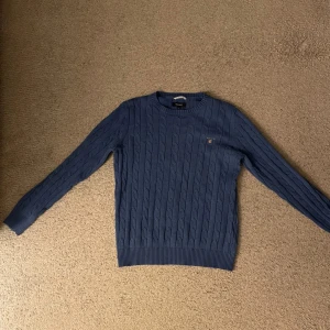 Blå kabelstickad tröja från GANT - Säljer en snygg blå kabelstickad tröja från GANT. Tröjan har en klassisk design med rund hals och klassiska Gant märket på framsidan. Hör av er vid funderingar!