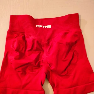 Röda DFYNE högmidjade träningsshorts - Säljer ett par röda högmidjade träningsshorts från DFYNE. De har en bred ribbad midja och är tillverkade i stretchigt material med 90% nylon och 10% elastan. Perfekta för gymmet eller andra sportaktiviteter. Snygg logga framtill och på sidan av midjan.