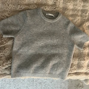 Grå stickad kortärmad tröja från Zara - Syns inte att den är använd. Supermjuk grå stickad tröja från Zara med korta ärmar och rund hals. Tröjan har en enkel och clean look med ribbade kanter vid ärmslut och nederkant. Perfekt till lager-på-lager eller bara som den är. 🥰🥰