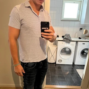 Randig pikétröja från Massimo Dutti - Säljer en stilren pikétröja från Massimo Dutti i vitt och marinblått. Tröjan har smala horisontella ränder, klassisk krage och korta ärmar. Materialet är mjuk bomull som känns skönt mot huden. Perfekt för dig som gillar en clean och fräsch look.