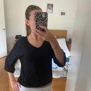 Svart v-ringad blus med trekvartsärm - Säljer en svart blus med v-ringning och trekvartsärmar. Toppen har en loose passform och är croppad vid midjan. Perfekt att styla med jeans eller kjol för en chill look. Materialet känns mjukt och lätt mot huden.