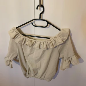 Beige offshoulder blus med volang - Supersöt beige offshoulder blus från Pee Ka Nong med bred volang runt halsringningen och volangdetaljer på ärmarna. Blusen har korta ärmar och resår i nederkanten för en snygg passform. Perfekt för dig som gillar romantisk och trendig stil.