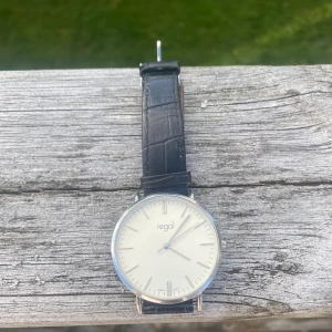Regal silverklocka med svart läderband - Stilren klocka från Regal med rund silverfärgad boett och minimalistisk vit urtavla. Svart band i läderimitation med krokodilpräglad yta och klassiskt spänne. Perfekt för dig som gillar enkel och clean design. Boetten är i rostfritt stål och klockan har japanskt urverk. Batteriet måste bytas.