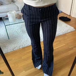 Mörkblå randiga bootcut kostymbyxor - Snygga mörkblå byxor med vita vertikala ränder och bootcut passform. Byxorna har hög midja, slits nertill på benen och klassiska fickor både fram och bak. Perfekta för dig som gillar en trendig och stilren look.