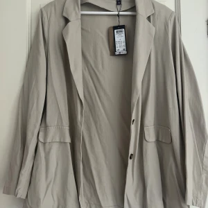 Beige kavaj från Vero moda - En helt ny  beige kavaj från Vero moda med klassisk krage och två framfickor med lock. Kavajen har två knappar framtill och är tillverkad i mjuk bomull. Perfekt för dig som gillar en stilren och enkel look.