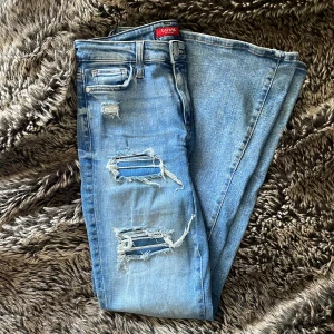 Blå bootcut jeans från Guess - Snygga blå bootcut jeans från Guess med slitna detaljer och hål på låren och knäna. Självsydd lågmidjade. Klassisk femficksmodell med dragkedja och knapp. Jeansen har en ljus tvätt och är tillverkade i jeansmaterial med lite stretch för skön passform.