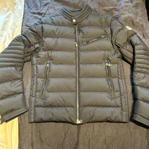Svart Moncler AUTHIE GIUBBOTTO - Använd fåtal gånger. Köpt på nk.Säljer en svart pufferjacka från Moncler med klassisk quiltad design och ståkrage. Jackan har tre dragkedjefickor framtill, snygga detaljer vid ärmslut och ikonisk Moncler-logga på ärmen. Perfekt för kalla dagar och riktigt stilren look.