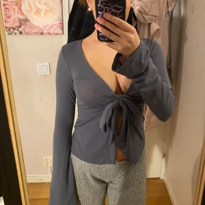 Grå långärmad knytblus Gina Tricot - Supersnygg och populär grå långärmad blus från Gina Tricot i storlek XS. Toppen har djup v-ringning och knyt framtill som ger en trendig look. Ärmarna är långa och lite utsvängda. Mjuk och stretchig i materialet, perfekt att styla med jeans eller kjol. Har för mig att denna modell e slutsåld. Endast testad!