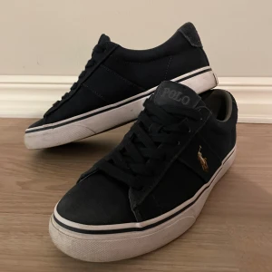 Ralph Lauren skor mörkblå - Mörkblåa sneakers från Polo Ralph Lauren med klassisk låg profil, vit sula och svart textilöverdel. Skorna har snörning, rund tå och den ikoniska Polo-loggan broderad i guld på sidan. Perfekta för en clean och stilren look.