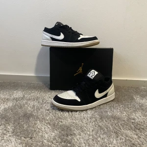 Nike Jordan 1 Low svart/vit - Snygga Nike Jordan 1 Low sneakers i svart och vitt med klassisk Swoosh på sidan. Skorna har lågt skaft, snörning och en vit sula. Perfekta för dig som gillar streetstyle och vill ha en ikonisk look.