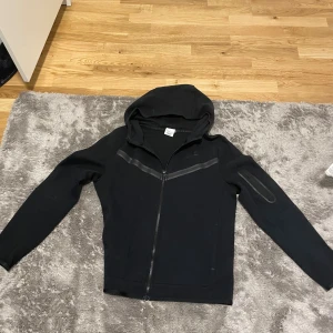 Svart Nike hoodie med dragkedja XL - Svart hoodie från Nike i storlek XL med hel dragkedja framtill och huva. Snygga detaljer med svarta ränder över bröstet och en extra ficka med dragkedja på ena ärmen. Perfekt för en sportig och avslappnad stil.