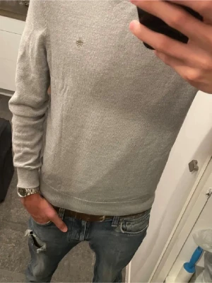 Grå stickad tröja från Morris - Snygg grå stickad tröja från Morris med diskret logga broderad på bröstet. Tröjan har rund hals, ribbade muddar och är långärmad. Perfekt att matcha med jeans eller chinos för en clean look.