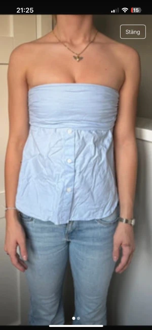 Ljusblå bandeau tubtopp med knappar - Säljer en ljusblå bandeau tubtopp med knappar framtill. Toppen är axelbandslös och har en enkel, rak form med lätt rynk upptill. Perfekt till jeans och sommarhäng. Materialet känns som bomull och toppen är ärmlös.