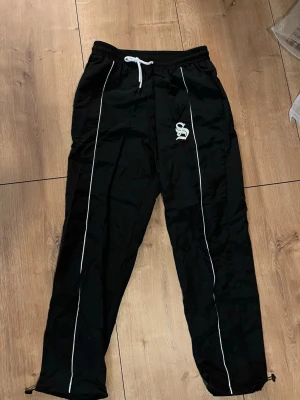 Svarta täckbyxor med vita detaljer - Svarta täckbyxor med vita ränder längs benen och vit snörning i midjan. Byxorna har elastisk midja och justerbara benslut med dragsko. Materialet är syntet och byxorna har en loose passform. Perfekt för dig som gillar streetwear. Helt nya , oanvända.