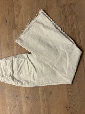 Vita jeans med rå kant och dubbelknapp - Snygga vita jeans med rak passform och rå, fransig kant längst ner på benen. De har två knappar i midjan och klassiska fickor både fram och bak. Materialet är jeans och färgen är ljusvit, vilket ger en fräsch vibe. Perfekt för dig som gillar en clean och trendig look.