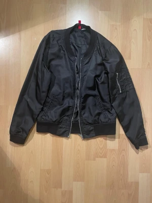 Svart bomberjacka från H&M - Svart bomberjacka från H&M. Jackan har klassisk ribbad krage, muddar och nederkant, samt en ficka med dragkedja på ärmen.
