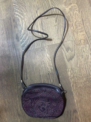 Brun axelväska med paisleymönster - Säljer en oval brun axelväska med paisleymönstrad framsida i mörka toner. Väskan har ett långt smalt axelband och dragkedja upptill. Materialet är en mix av skinn och textil, insidan är fodrad i vinrött tyg. Perfekt storlek för mobilen och småsaker. Tyvärr behövs en händighand för att rädda denna