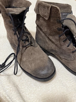 Bruna mockakängor från Collusion - Säljer ett par bruna kängor från Collusion Denham med grov mocka och svarta snören. Skorna har rund tå, platt sula och högt skaft med breda tungor. Perfekt för dig som gillar en robust och cool stil. Märke syns tydligt på tungan.