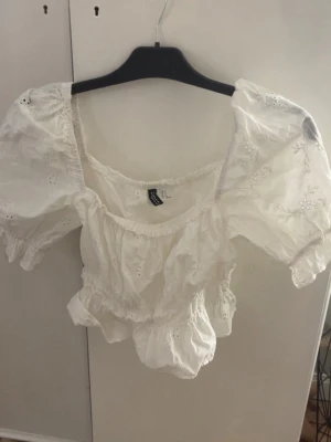 Vit blus från H&M Divided - Supersöt vit blus från H&M Divided med puffärmar och broderade blommor. Modellen är croppad med resår i midjan och vid ärmsluten. Perfekt till sommarens alla outfits och har en romantisk vibe med volangdetaljer och fyrkantig ringning.