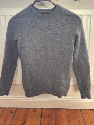 Grå stickad tröja från Morris - Säljer en grå stickad tröja från Morris med rund hals och diskret blå logga broderad på bröstet. Tröjan har långärmade ribbade muddar och är gjord i mjukt ullmaterial, perfekt för kyliga dagar.