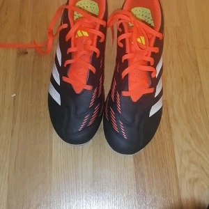 Adidas Predator fotbollsskor svart/röd - Säljer ett par Adidas Predator fotbollsskor med svart ovandel, röda snören och detaljer samt vita ränder på sidan. Skorna har platt sula med dobbar för gräsplan och är gjorda i syntetmaterial. Snygg design med röda grafiska mönster på sidorna och klassisk Adidas-logga.