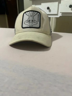Beige och grå keps från Capslab - Säljer en beige och grå keps från Capslab med Bugs Bunny-tryck framtill. Kepsen har mesh på sidorna och bak, justerbar snapback och texten 'That's all Folks!' broderad på sidan. Perfekt för dig som gillar Looney Tunes och vill ha en unik accessoar.