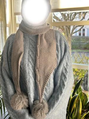 Grå stickad väst med luva och tofsar - Säljer en grå stickad väst med luva och långa snören som avslutas med fluffiga tofsar. Västen har en mysig känsla och är perfekt för kalla dagar. Den är ribbstickad och har en enkel, stilren design som passar till många outfits.