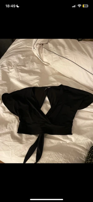 Svart omlottopp från H&M Divided - Säljer en svart omlottopp från H&M Divided i storlek M. Toppen har korta, vida ärmar och knyt framtill i midjan. Materialet är mjukt och stretchigt, perfekt för att skapa en cool och avslappnad look. Passformen är croppad och toppen har en djup v-ringning.