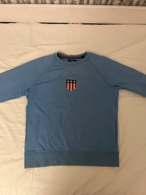 Ljusblå sweatshirt från GANT - Ljusblå sweatshirt från GANT med broderad logga och sköld på bröstet. Klassisk rund halsringning och långa ärmar. Tillverkad i mjuk bomull, perfekt för chill dagar. Snygg detalj med röd, vit och blå sköld som ger en sportig vibe.