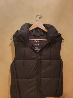 Svart dunväst från Zara med huva - Säljer en svart dunväst från Zara med stor, mysig huva och dragkedja framtill. Västen har två sidofickor med dragkedja och justerbara snören vid huvan. Materialet är syntet och polyester, perfekt för kyliga dagar. Stilren och enkel design.