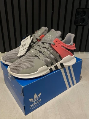 Adidas EQT Support ADV sneakers grå/rosa - Säljer ett par Adidas EQT Support ADV sneakers i grå mesh med detaljer i ljusgrått och svart samt en snygg rosa plastdetalj vid hälen. Skorna har snörning, platt sula och klassiska tre svarta ränder på sidan. Perfekt för dig som gillar sportig och modern stil.